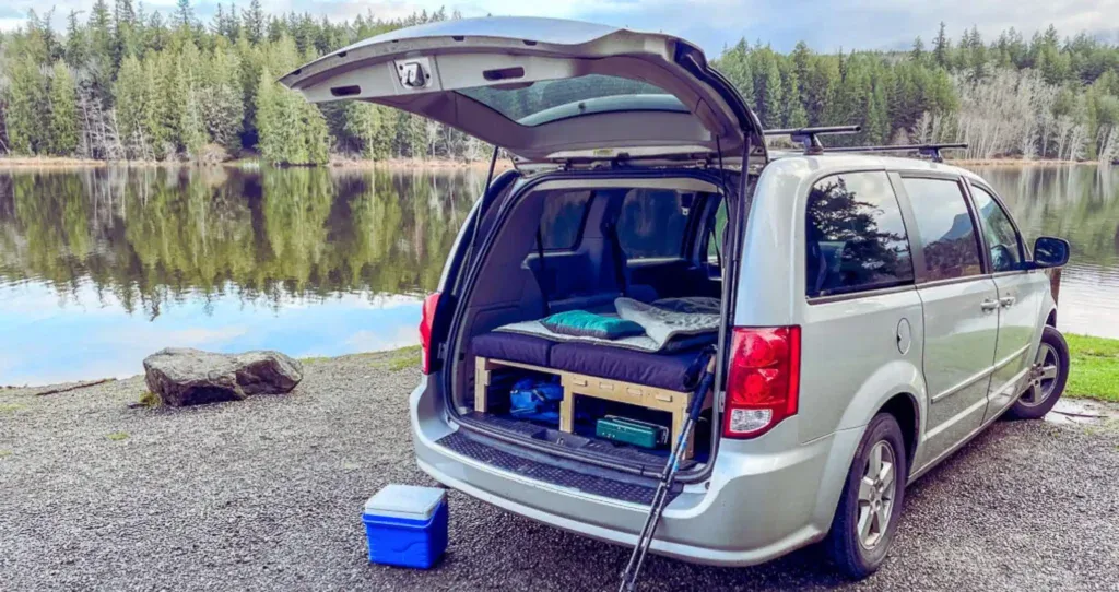 Coche camper equipado para acampar junto a un lago, con tienda plegada en el maletero y ambiente natural de bosques y agua. Ideal para aventuras y viajes en vehículo homologado.