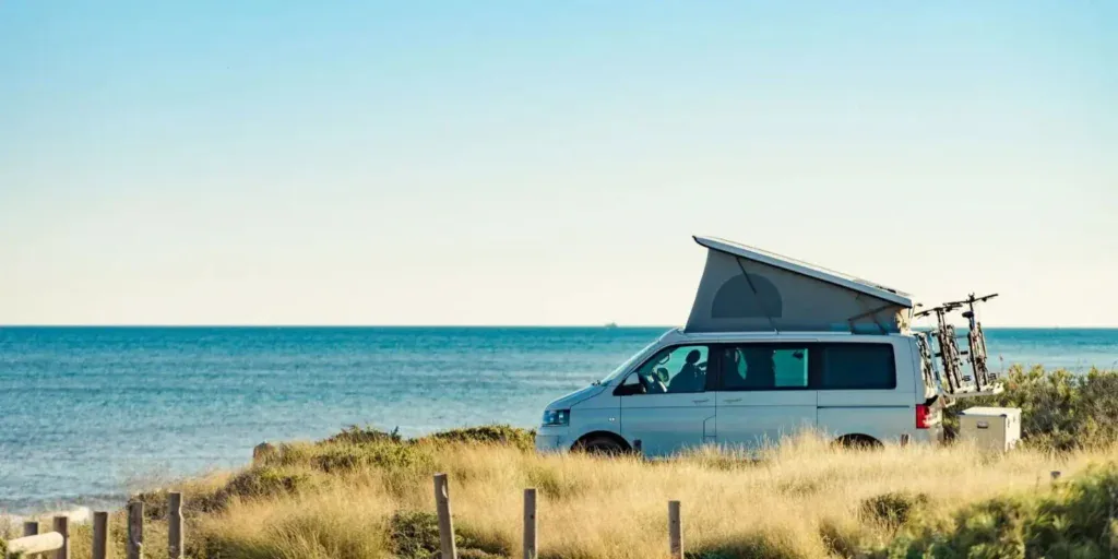 ALT: Camper equipado para viajes por la playa con cielo despejado, bicicletas en la parte trasera, y techo elevable, listo para explorar y disfrutar de la naturaleza y el mar.