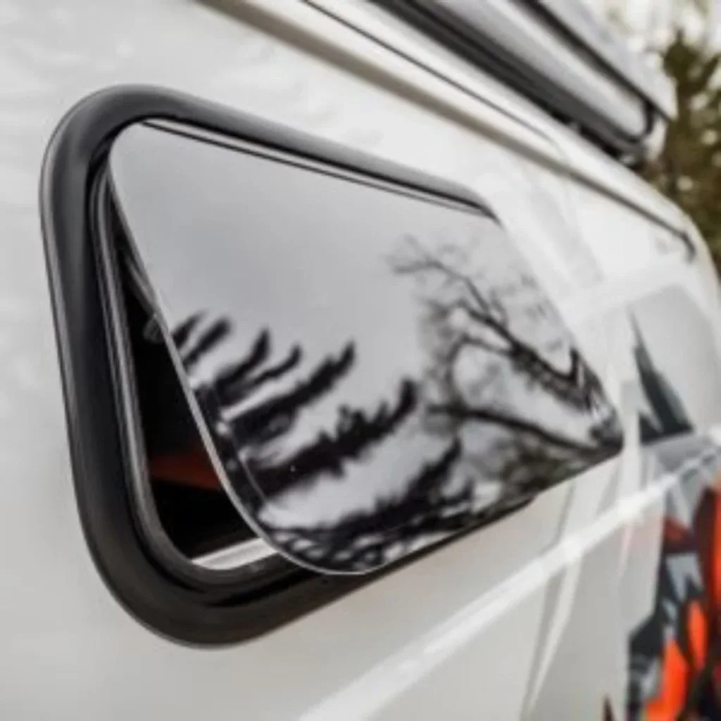 Ventana de camper van con cierre y reflejo de árboles en el cristal, ideal para conversiones de vehículos y homologaciones.